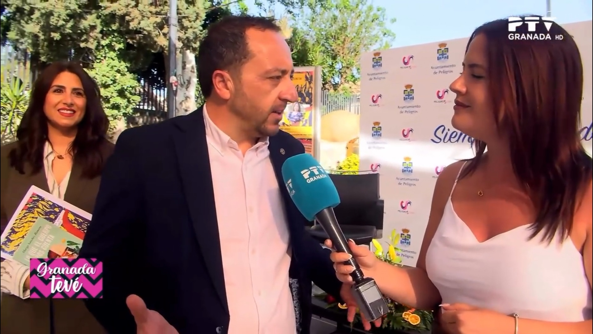 Laura Nieto, periodista de PTV Granada, entrevista al alcalde de Peligros, Roberto García, durante la inauguración de la XIV Feria del Libro, en presencia de la concejala Cristina López Núñez al fondo.